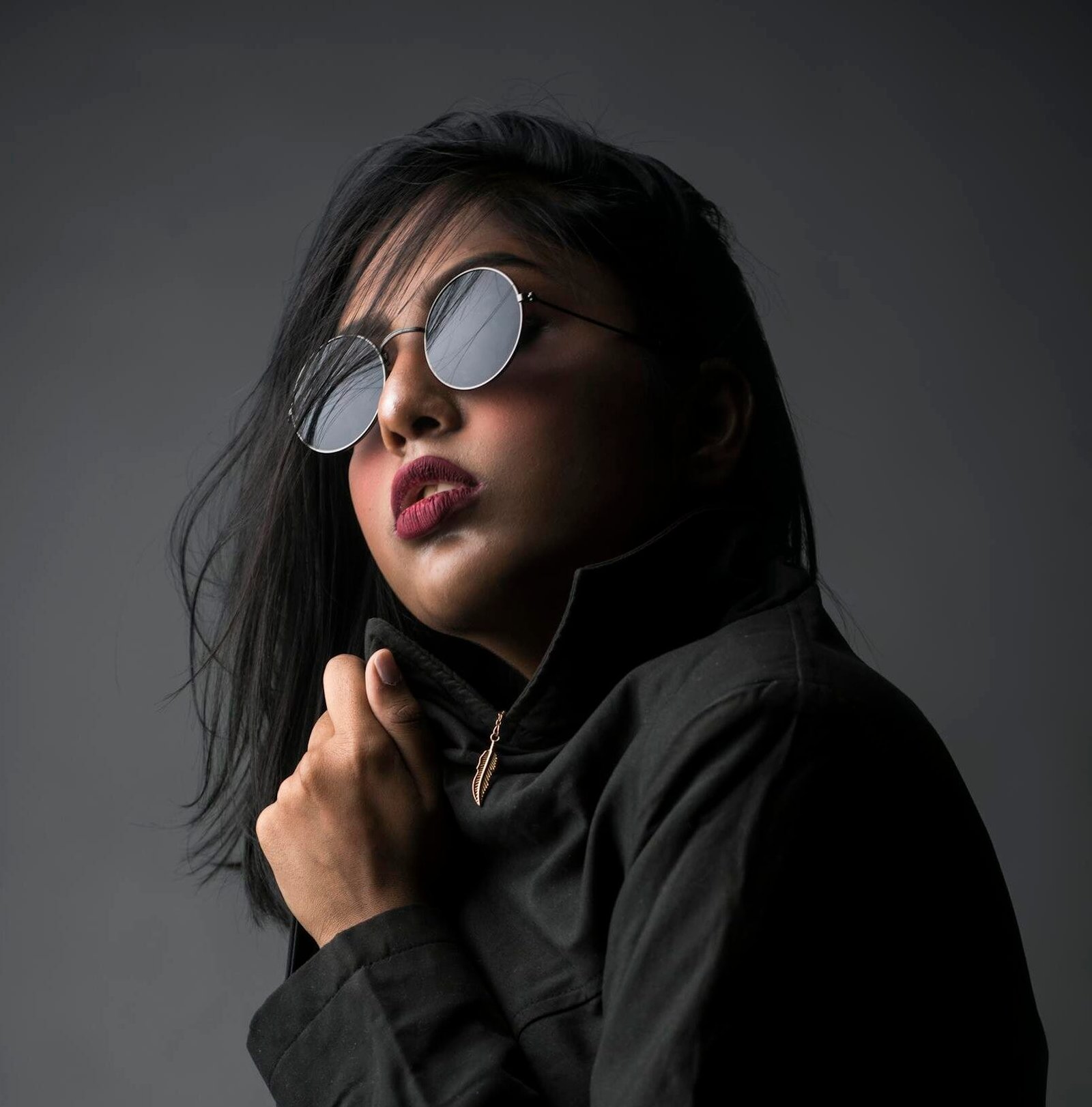 Stylish portrait of a woman posing in a studio with dark fashion attire and round sunglasses.